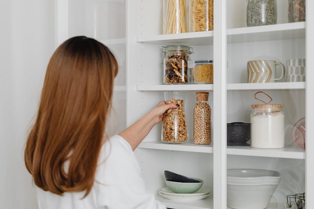 Woman organizing your new pantry by arranging jars of pasta, grains, and dried fruits on white shelves